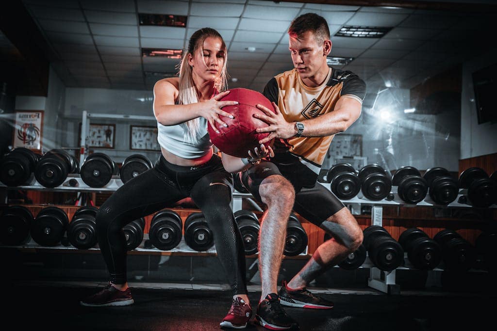 A man and woman perform a tandem workout with a medicine ball in a gym setting.